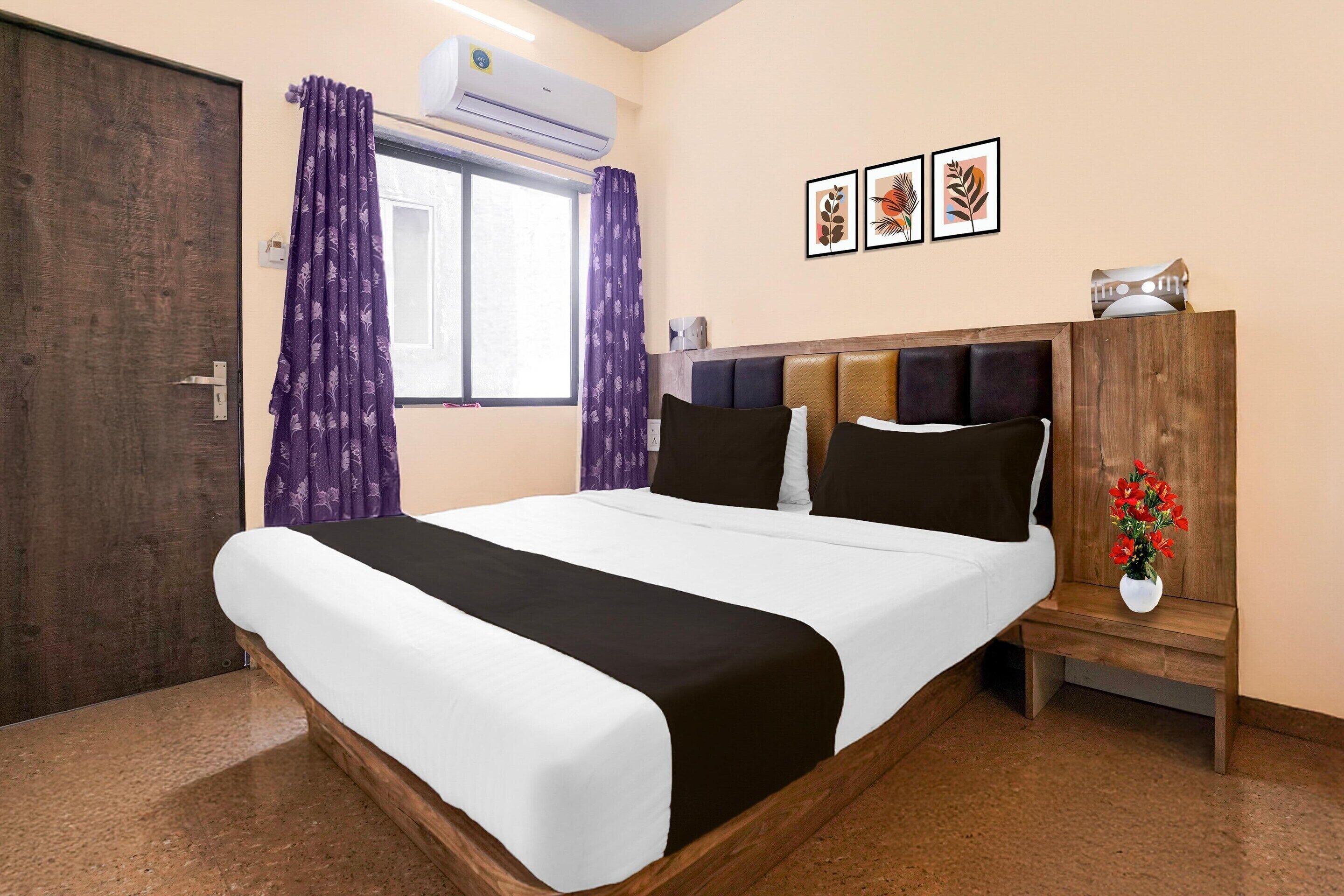 A hotel room with a bed, a nightstand, a window with curtains, an air conditioning unit, and framed artwork on the wall.