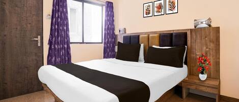 A hotel room with a bed, a nightstand, a window with curtains, an air conditioning unit, and framed artwork on the wall.