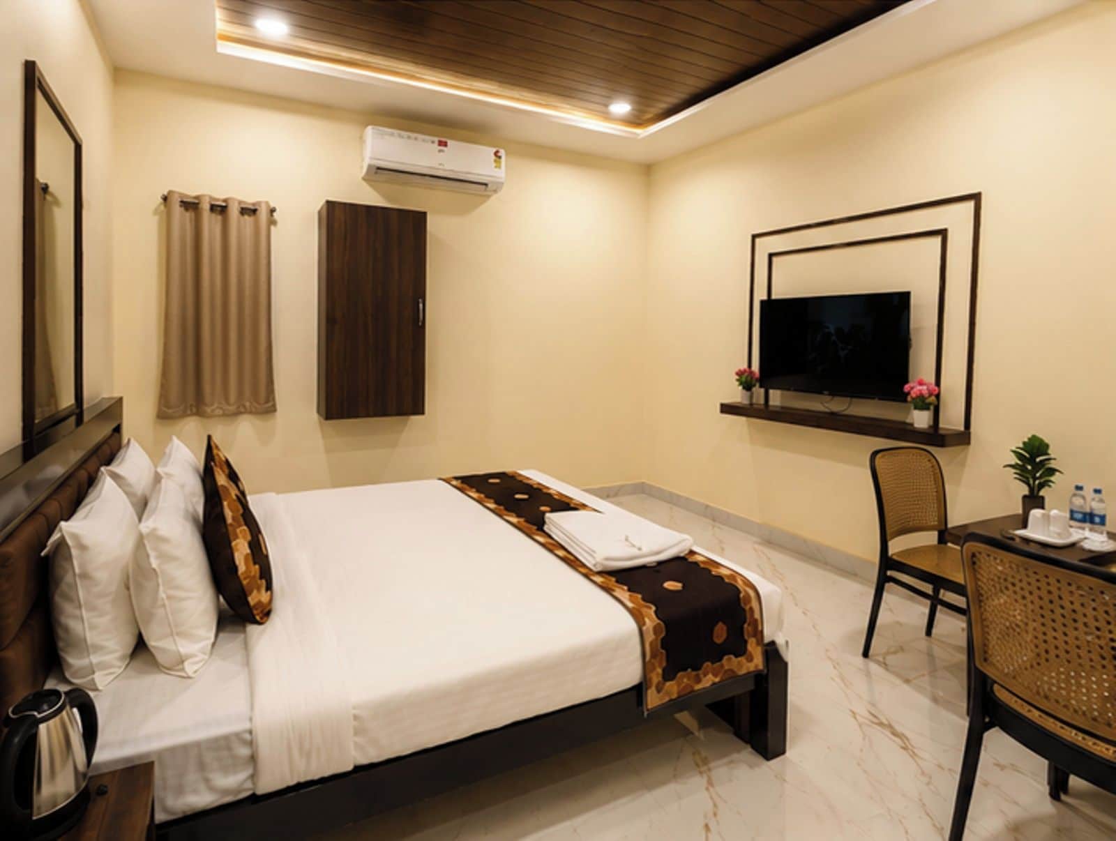 A hotel room with a bed, a television mounted on the wall, a chair, and a table with a kettle and water bottles.
