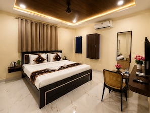 A hotel room with a bed, a desk, a chair, a wardrobe, a mirror, and an air conditioning unit.