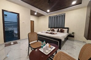 Una habitación de hotel con una cama, una mesa de comedor, sillas, una mesita con una bandeja de objetos, una ventana con cortinas y una puerta que da a un balcón.