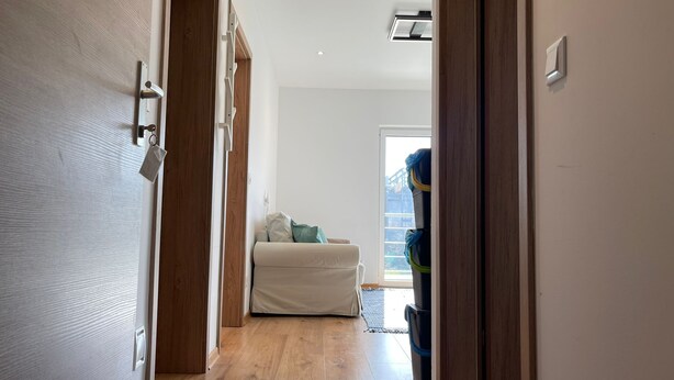 Un pasillo con una puerta de madera, una pared blanca, una lámpara en el techo y vista a una habitación con cama y ventana.