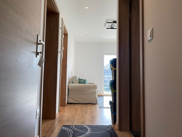 A hallway with a wooden door, a white wall, a ceiling light, and a view of a room with a bed and a window. - Cozy Apartment near Vienna Airport # 2 /free Parking/Self Check-in (Schwechat)