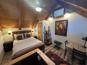 A bedroom with a wooden ceiling, a bed with pillows, a desk with a chair, a bookshelf, and a painting on the wall.