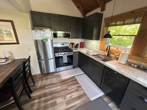 A modern kitchen with dark cabinets, stainless steel appliances, a wooden dining table, and a window with a view of greenery.