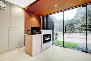 Un comptoir d’accueil avec une enseigne indiquant qu’il est géré par OYO, un intérieur moderne avec un plafond en bois et une vue sur la rue et la verdure environnante.