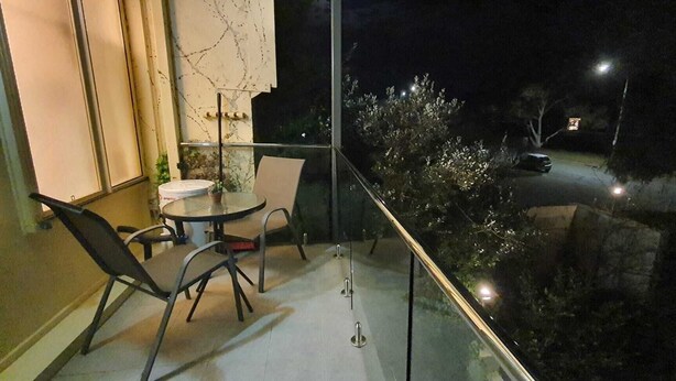 Een balkon met een glazen hekwerk, een ronde tafel, twee stoelen en een bloempot met een plant.