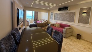 Un salon moderne doté d’un grand téléviseur à écran plat, d’une table à manger aux lignes illuminées, d’un ensemble de canapés et offrant une vue sur la mer grâce à une grande fenêtre.
