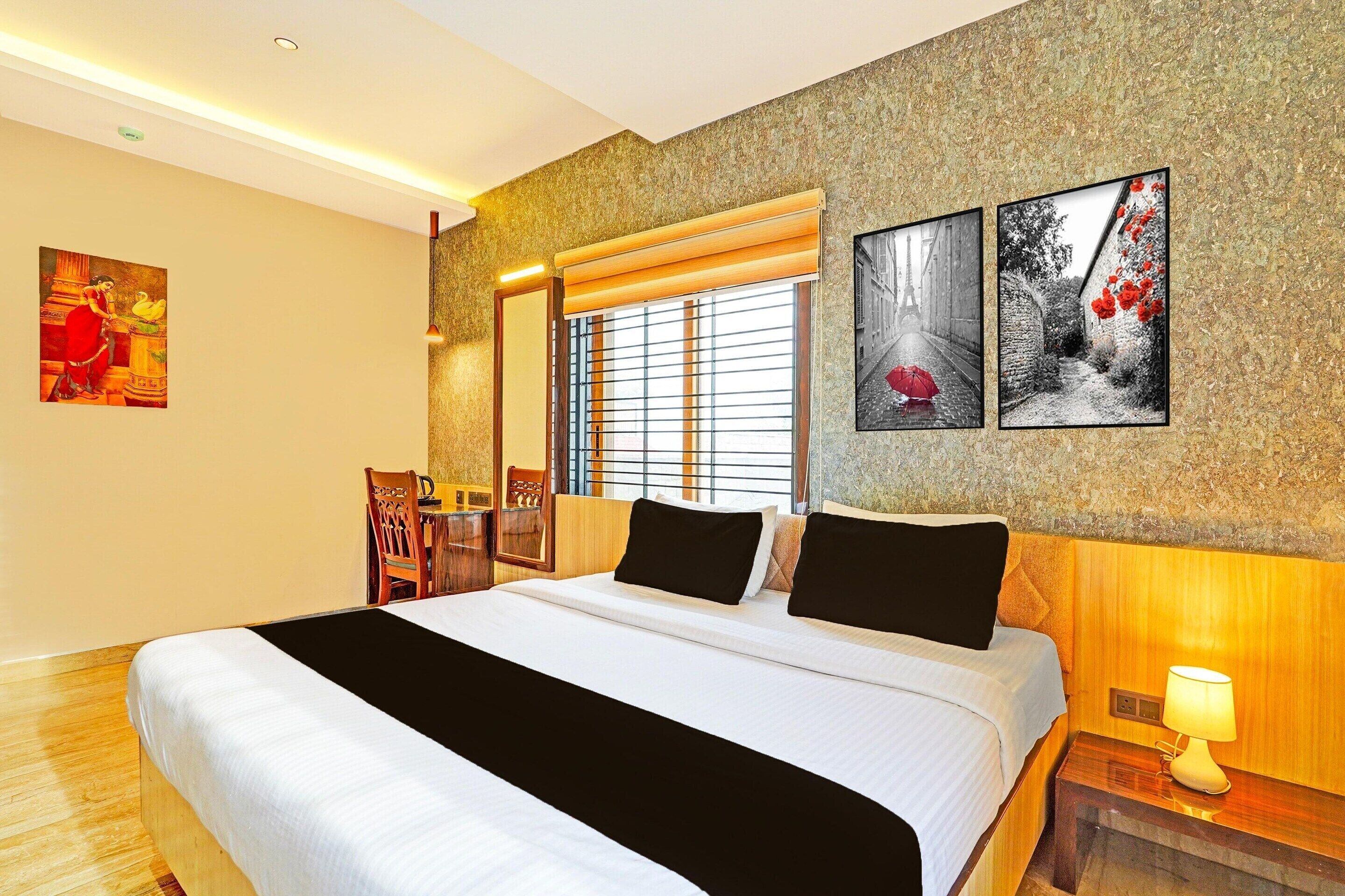 Habitación de hotel con una cama grande, una mesita de noche de madera con lámpara, una ventana con persianas y obras de arte en las paredes.
