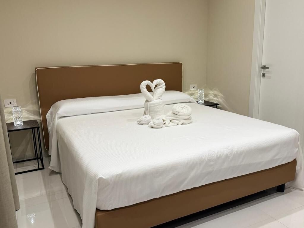 A neatly made bed with a heart-shaped towel and rolled towels on a white bedspread, a bedside table with a glass, and a door to the right.