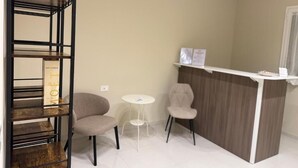 Zona de recepción con un mostrador de madera, dos sillas, una pequeña mesa blanca y una estantería.