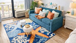 Une pièce avec un canapé bleu, un tapis orné de motifs d’étoiles de mer et une vue sur l’océan.