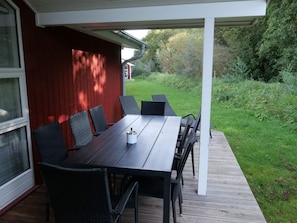 A covered outdoor dining area with a table and chairs, a wooden deck, and a red wall. - 4 Star Holiday Home in Grossenbrode (Grossenbrode)