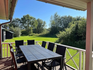 Ein überdachter Außenbereich mit Tisch und Stühlen, Blick auf ein grünes Feld und ein kleines Gebäude im Hintergrund.