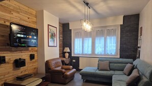 A living room with a television, wooden wall paneling, a brown leather chair, a green sofa, and a window with curtains.