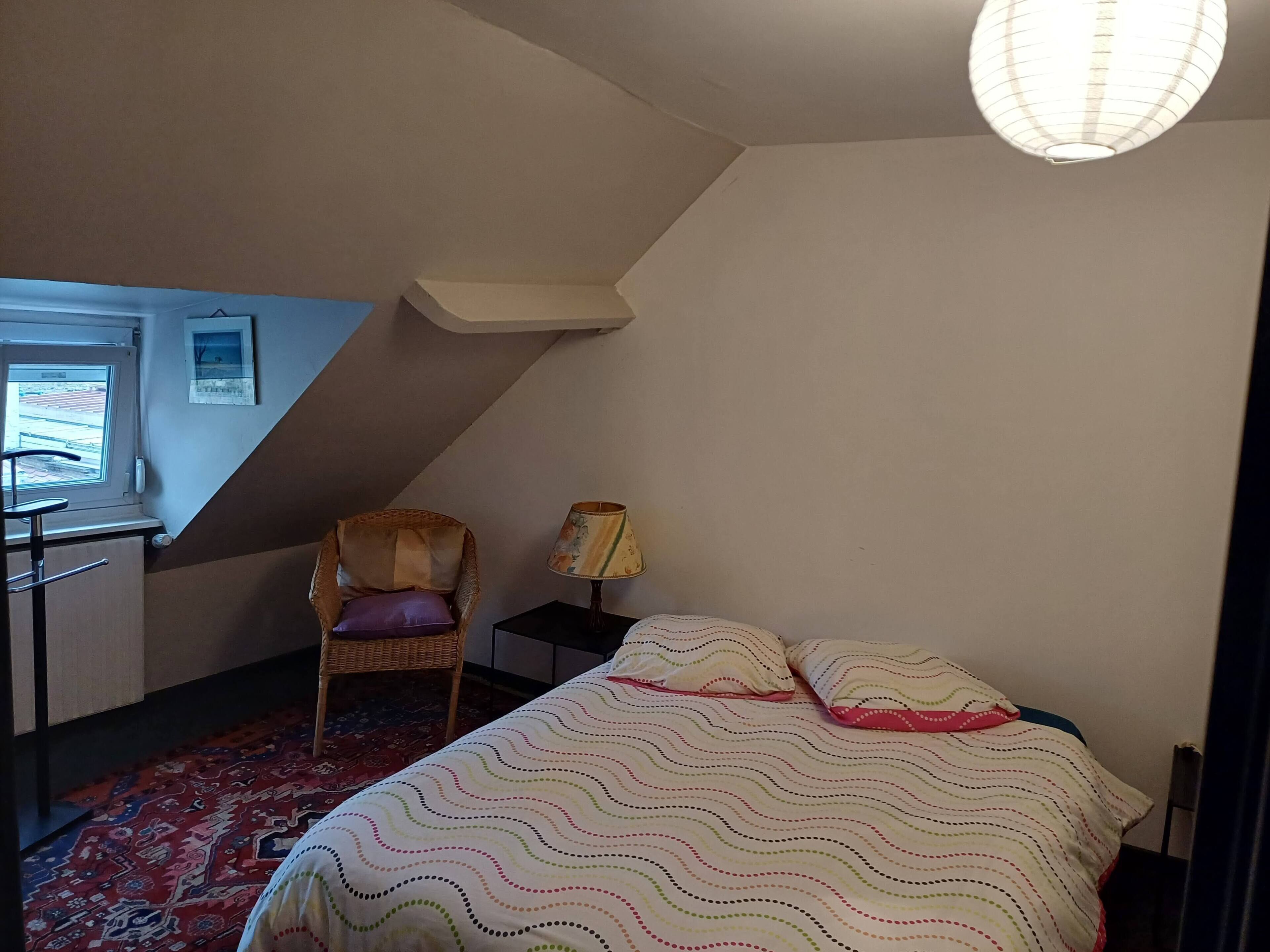 A bedroom with a bed, a chair, a lamp, a window, and a small table.