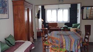 A room with a bed, a dining table with chairs, a wooden wardrobe, a wall clock, and a window with curtains.