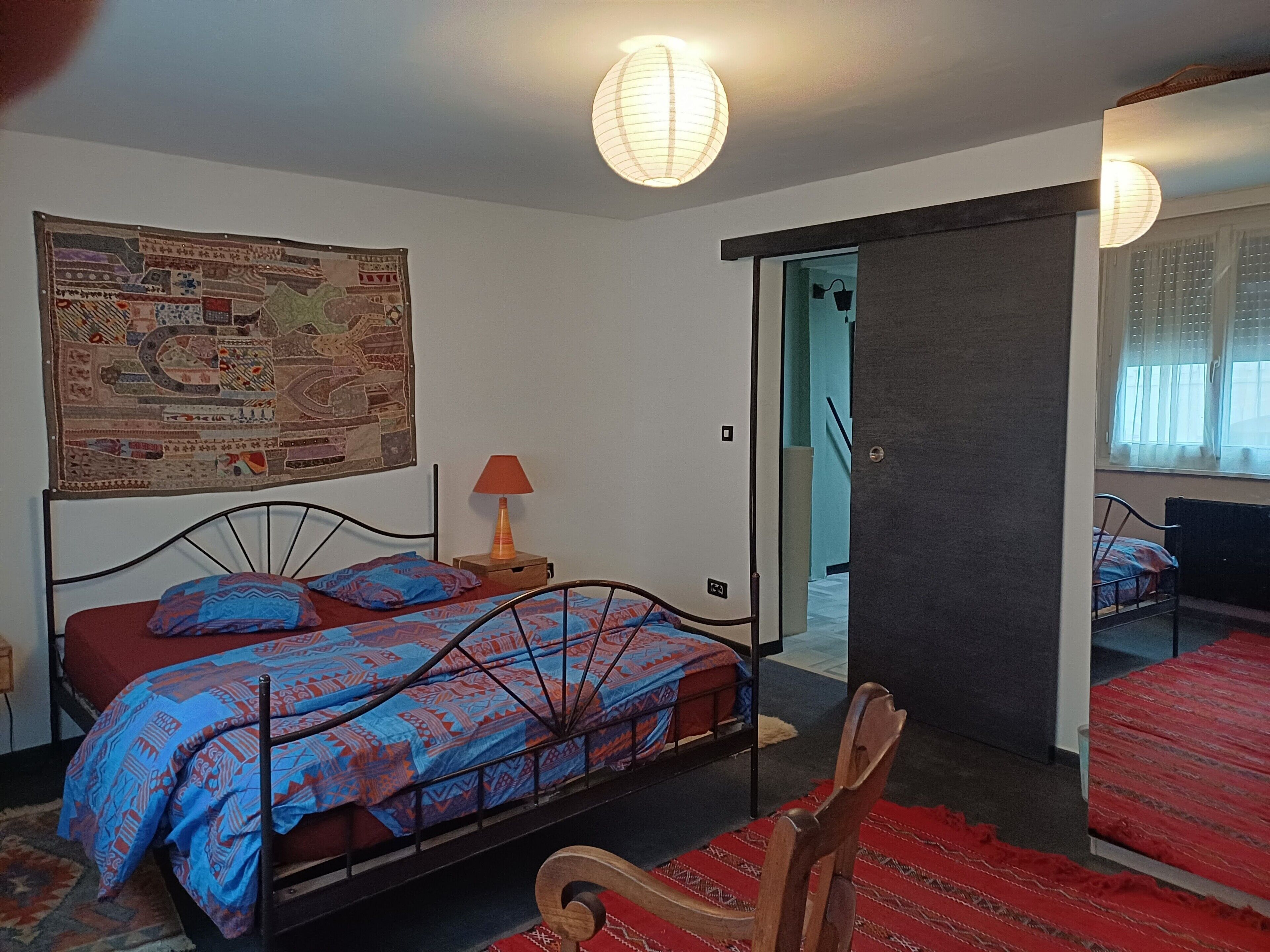 A bedroom with a bed, a nightstand, a lamp, a chair, a map on the wall, and a sliding door leading to another room.