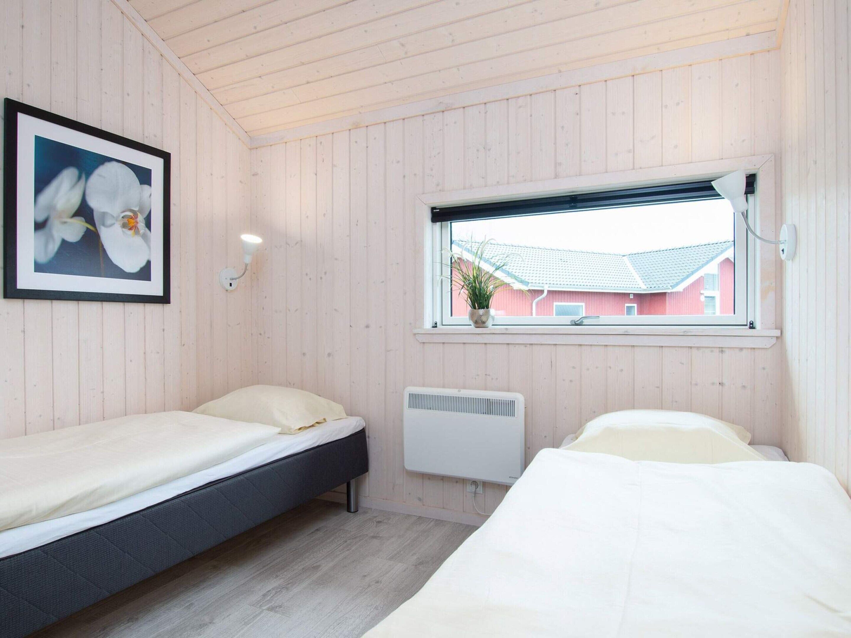 A room with two beds, a window with a view of a red building, a wall-mounted light, and a framed picture of a flower.