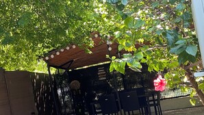 Un espacio de estar al aire libre con techo, sillas y una mesa, rodeado de follaje y una cerca.