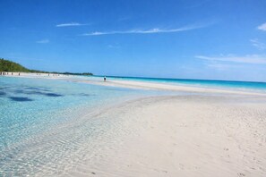 Une plage à l’eau turquoise et limpide, au sable blanc, bordée d’une zone boisée qui s’étend au loin.