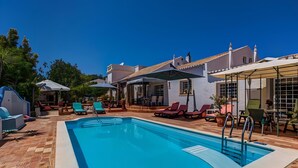 A swimming pool with lounge chairs and umbrellas, surrounded by a patio area with a tiled floor. - Algarve Apartment With Garden & Pool (Portimão)