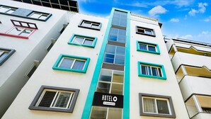 A hotel with a central vertical glass corridor, multiple windows with teal frames, and a sign indicating it is a company-serviced hotel. - Single Room Olivia Comforts (Bengaluru)