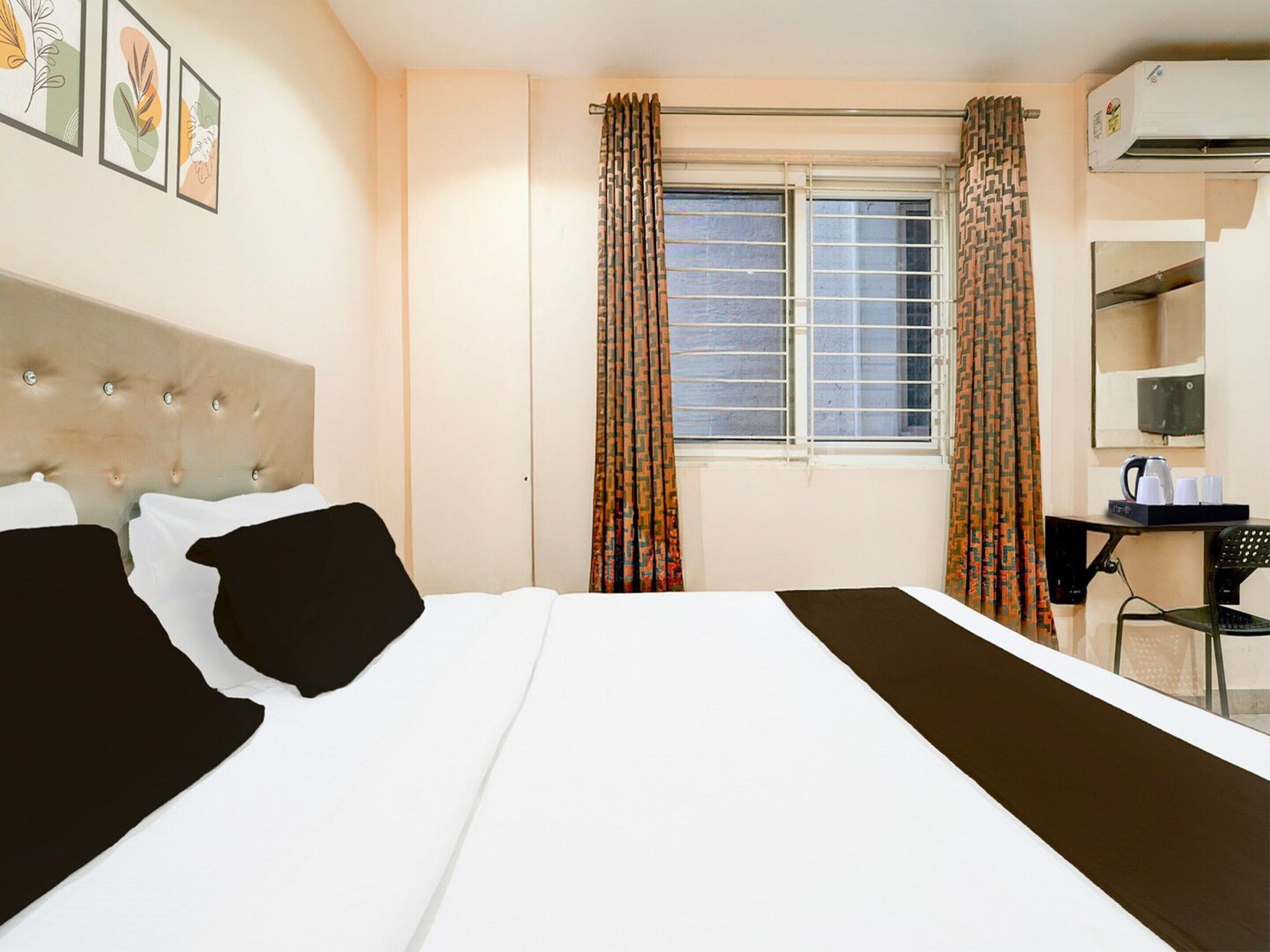 A hotel room with a bed, a desk with a chair, a window with curtains, and an air conditioning unit.