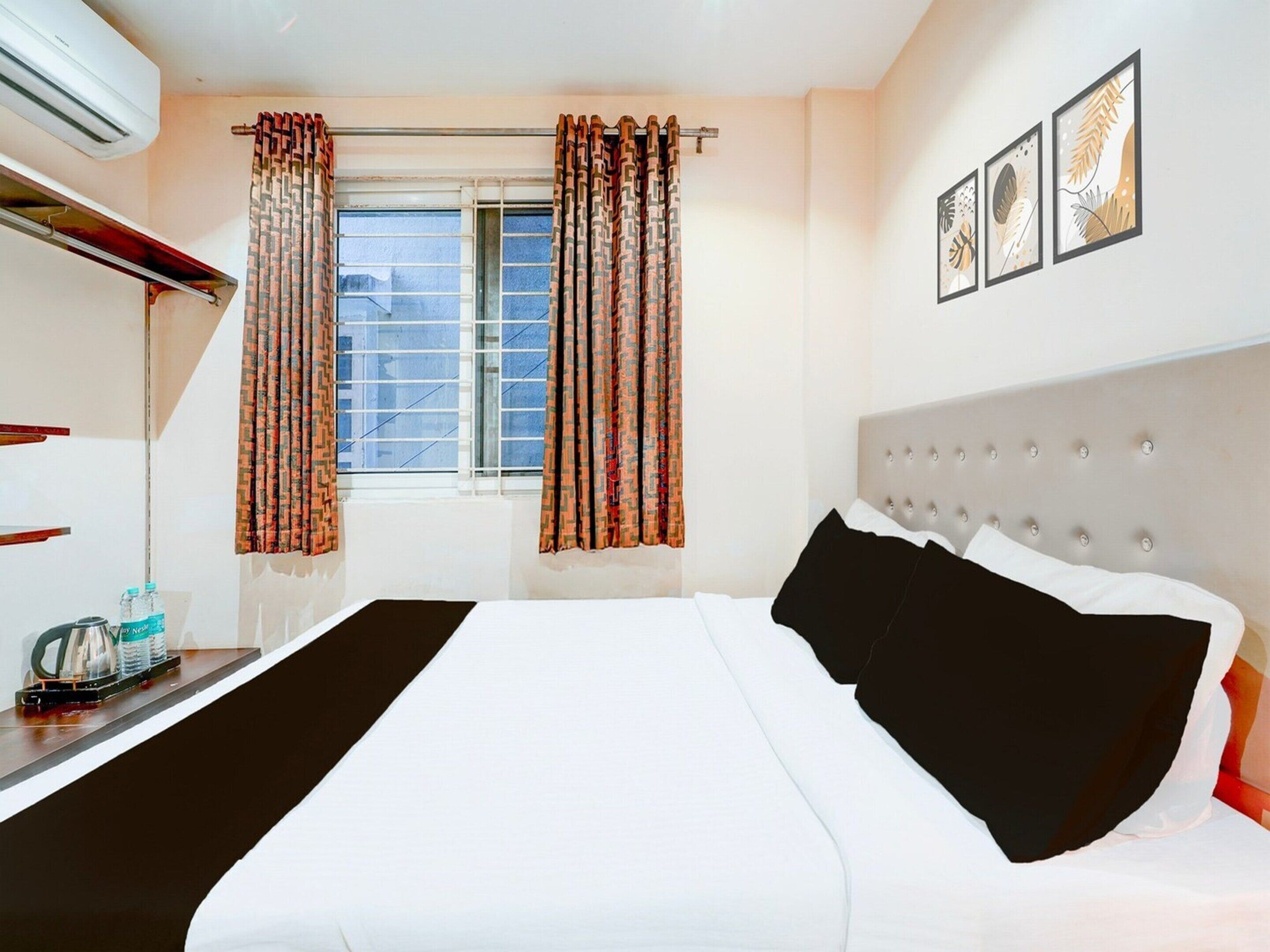 A hotel room with a bed, curtains, a window, a wall-mounted picture, and a bedside table with a kettle.