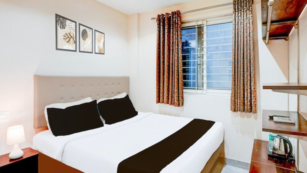 A hotel room with a bed, a nightstand, a lamp, a window with curtains, and a view of the city. - Single Room Olivia Comforts (Bengaluru)