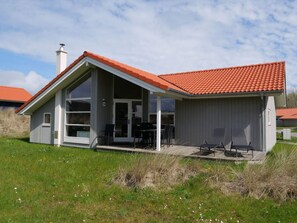 Et etplanshus med rødt tegltag, en overdækket terrasse med udendørs møbler og et stort vindue.