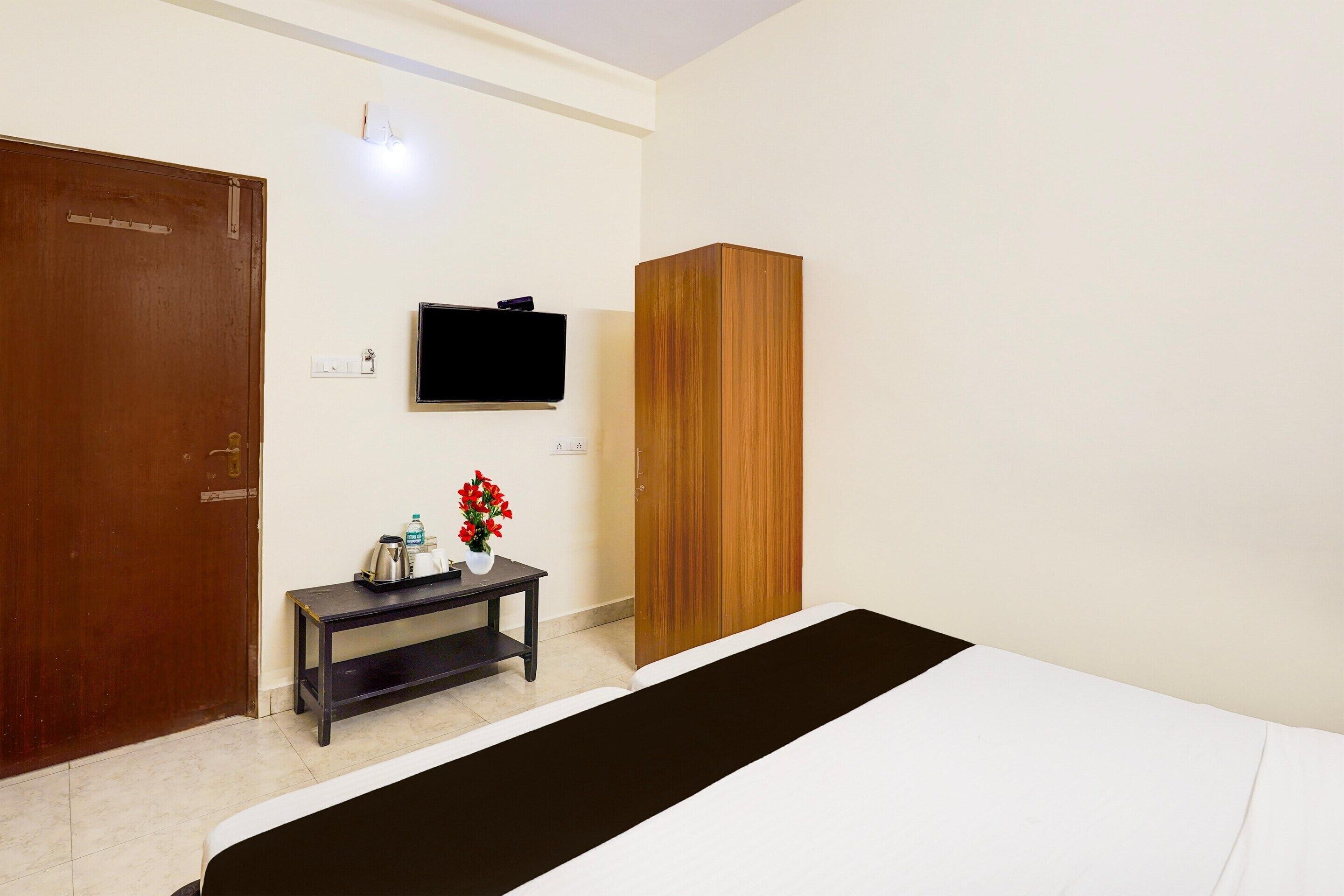 A hotel room with a bed, a wooden wardrobe, a small table with a vase of flowers, a television mounted on the wall, and a door.