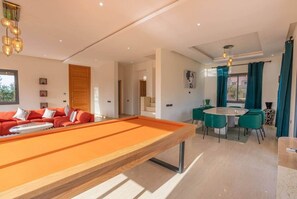A modern living room with a pool table, red sofa, and a dining area with teal chairs.