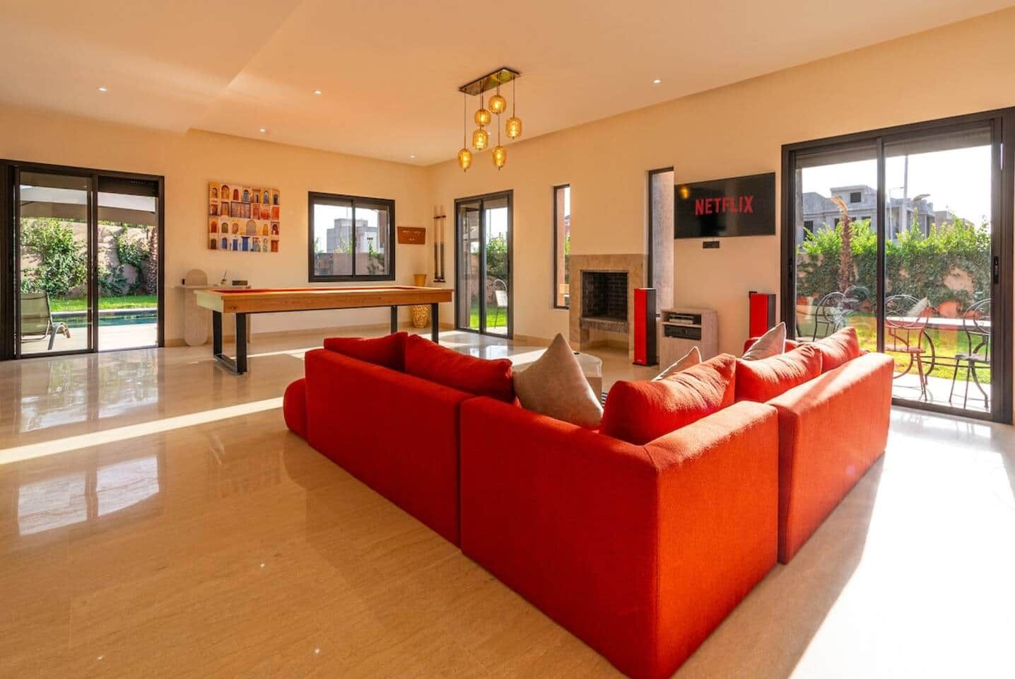 A modern living room with a red sofa, a wooden coffee table, a fireplace, and a large window with a view of greenery.