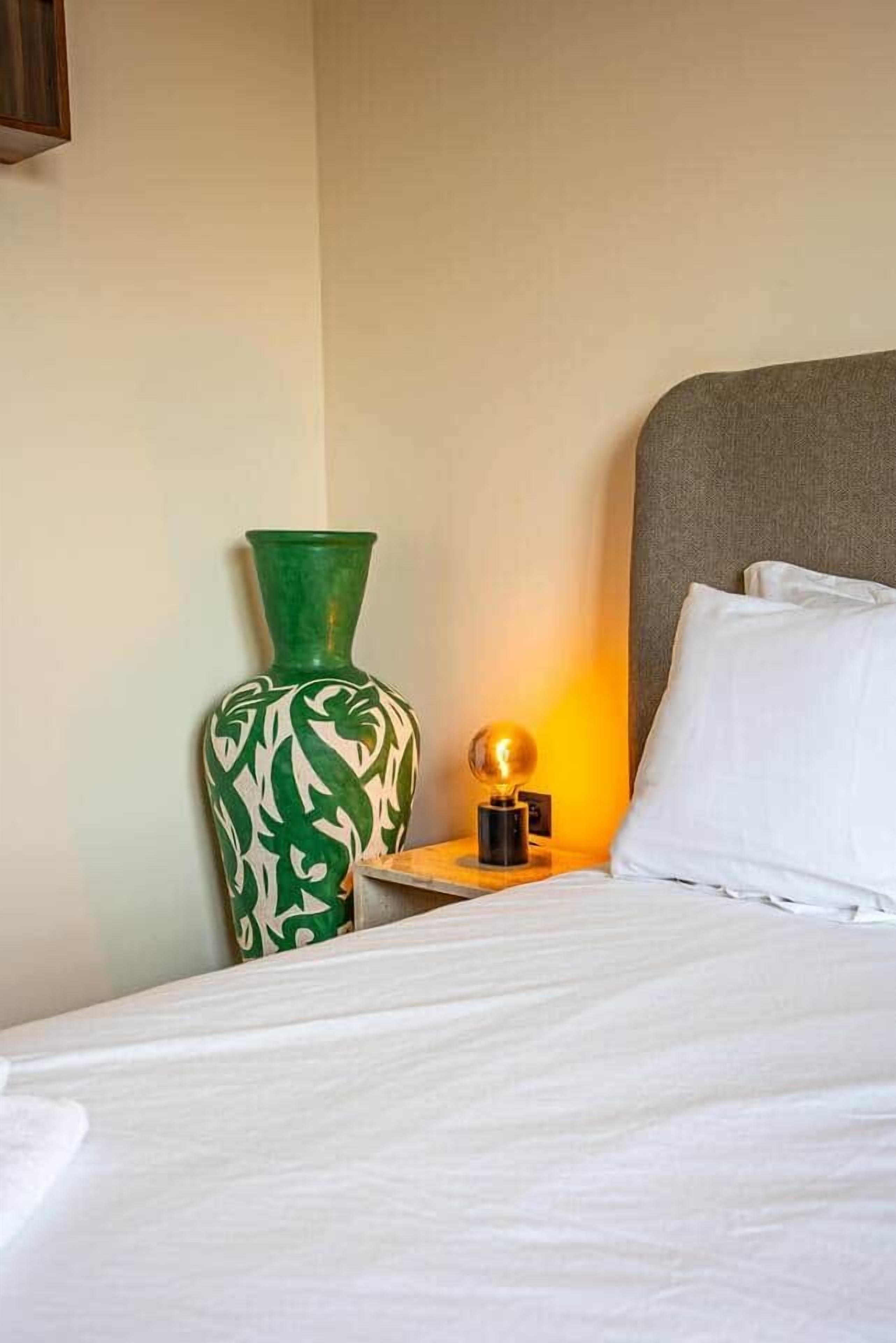 Une chambre d’hôtel avec un lit, des oreillers blancs, un vase vert et une petite table avec une lampe.