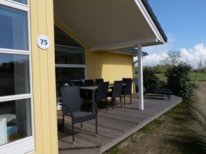 A covered outdoor area with a table and chairs, a number sign '75' on the wall, and a view of the surrounding landscape. - 4 Star Holiday Home in Grossenbrode (Grossenbrode)