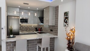 A modern kitchen with grey cabinets, a stainless steel refrigerator, a microwave, and a sink. There are three bar stools and a decorative plant. - Sunny Poolside Modern Apartment in Tenerife South (CN)