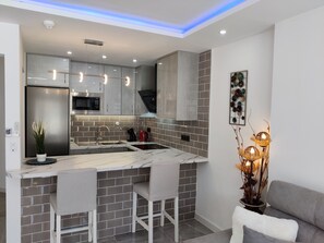 Eine moderne Küche mit grauen Schränken, einem Edelstahl-Kühlschrank, einer Mikrowelle und einem Spülbecken. Drei Barhocker und eine Zierpflanze sind ebenfalls vorhanden.