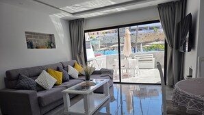 A modern living room with a grey sofa, a coffee table, and a view of the pool and outdoor seating area. - Sunny Poolside Modern Apartment in Tenerife South (CN)