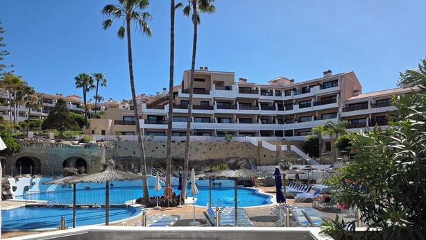 A hotel with a pool area, palm trees, and a stone wall. - Sunny Poolside Modern Apartment in Tenerife South (CN)