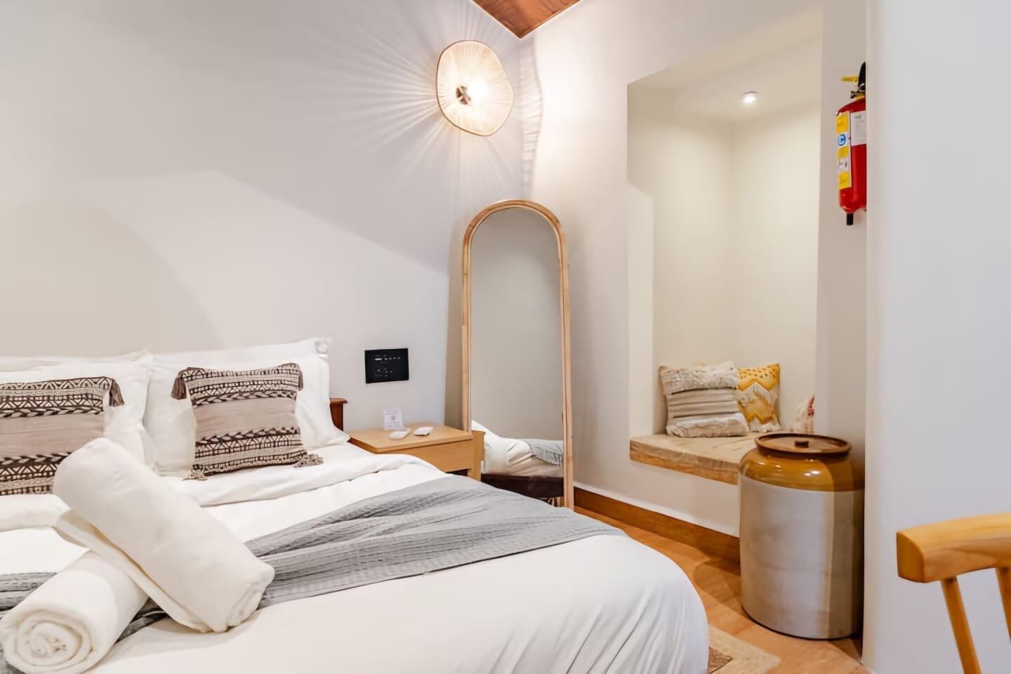 A hotel room with a bed, a bedside table, a mirror, a wall-mounted light, and a fire extinguisher.