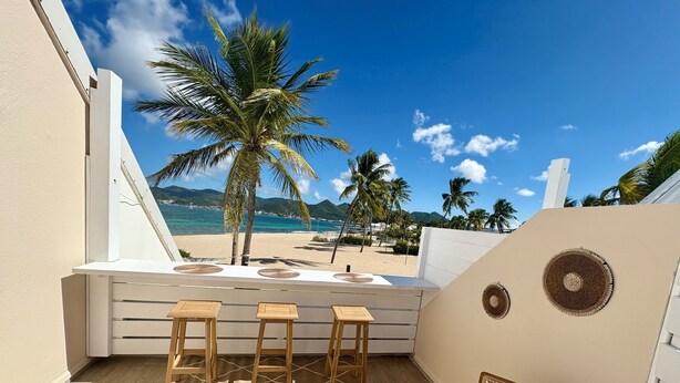 Un balcone con vista sulla spiaggia, sgabelli di legno, un tappeto intrecciato e un elemento decorativo a parete.