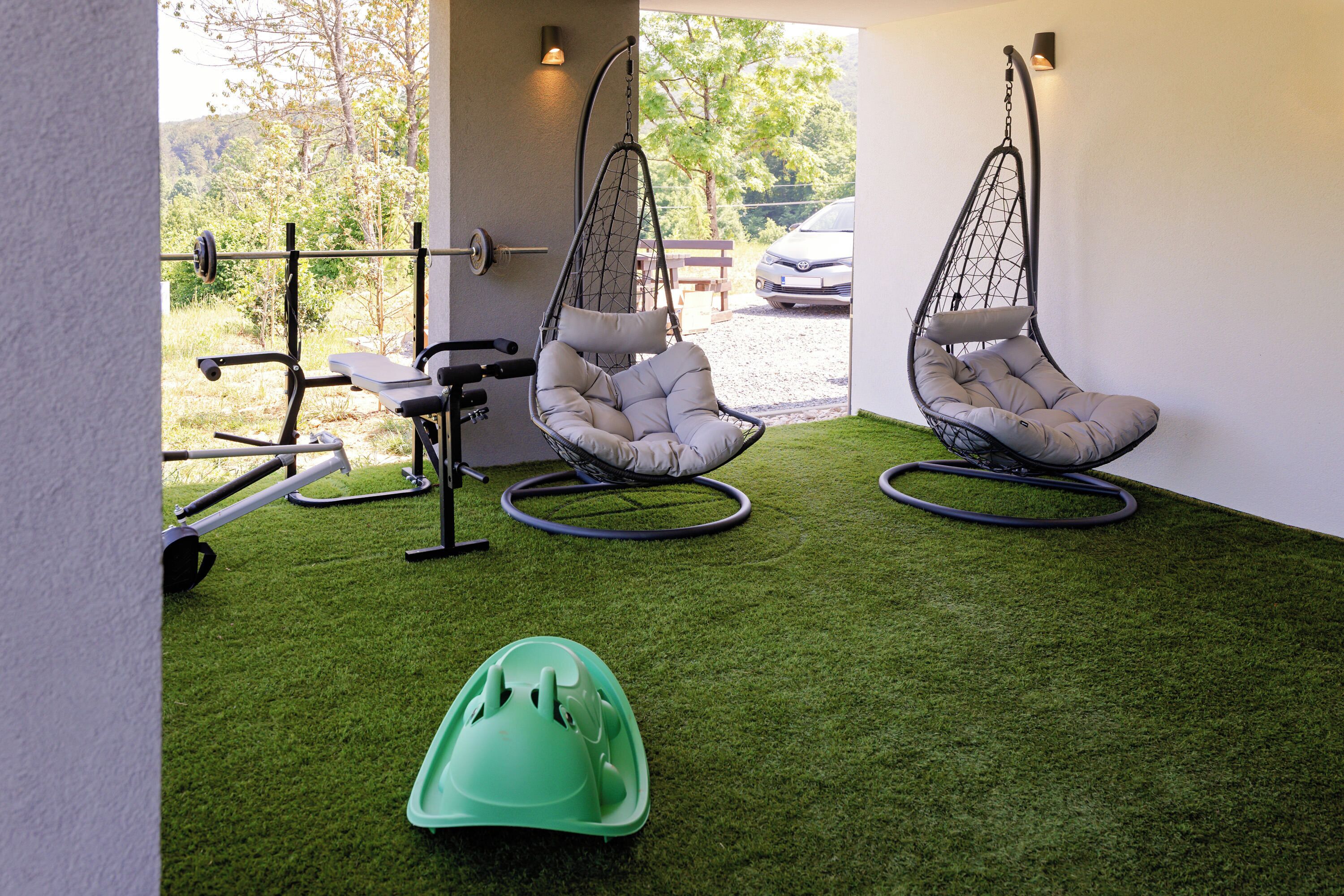 Un espace extérieur moderne comprenant deux fauteuils suspendus, un tapis de sol vert pour les exercices et un appareil de musculation.