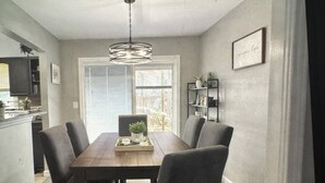Un coin repas avec une table en bois, six chaises, un lustre, une fenêtre et une cuisine en arrière-plan.