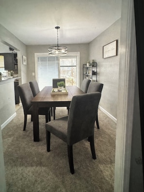 A dining area with a wooden table, six chairs, a chandelier, a window, and a kitchen in the background.