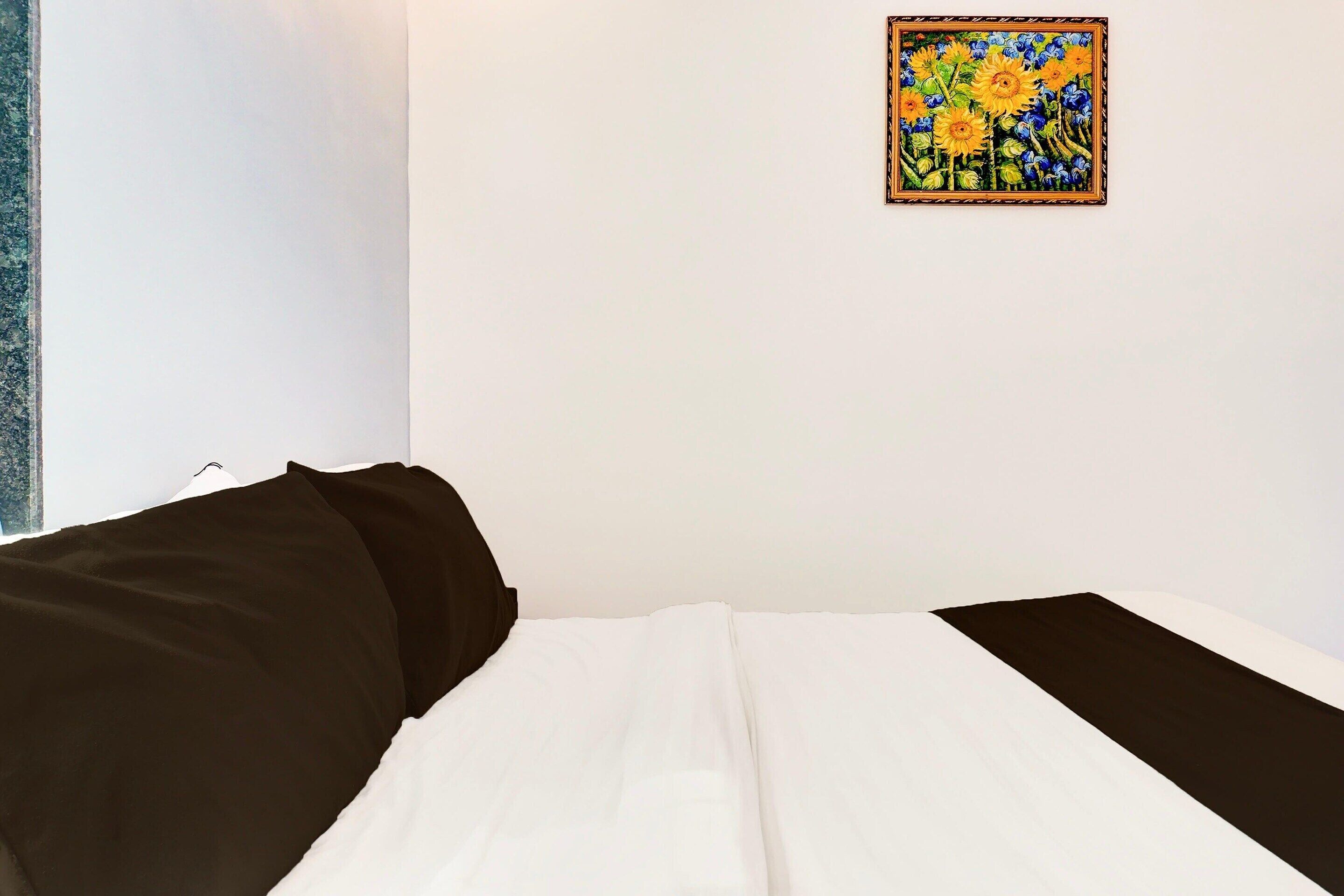 A bed with a white and brown bedspread, a painting of sunflowers on the wall, and a window with a view of the sky.