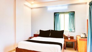 A bedroom with a wooden bed, a bedside table with a lamp, a window with curtains, and a small wooden cabinet. - Classic With Balcony Ubale Nagar (Pune)
