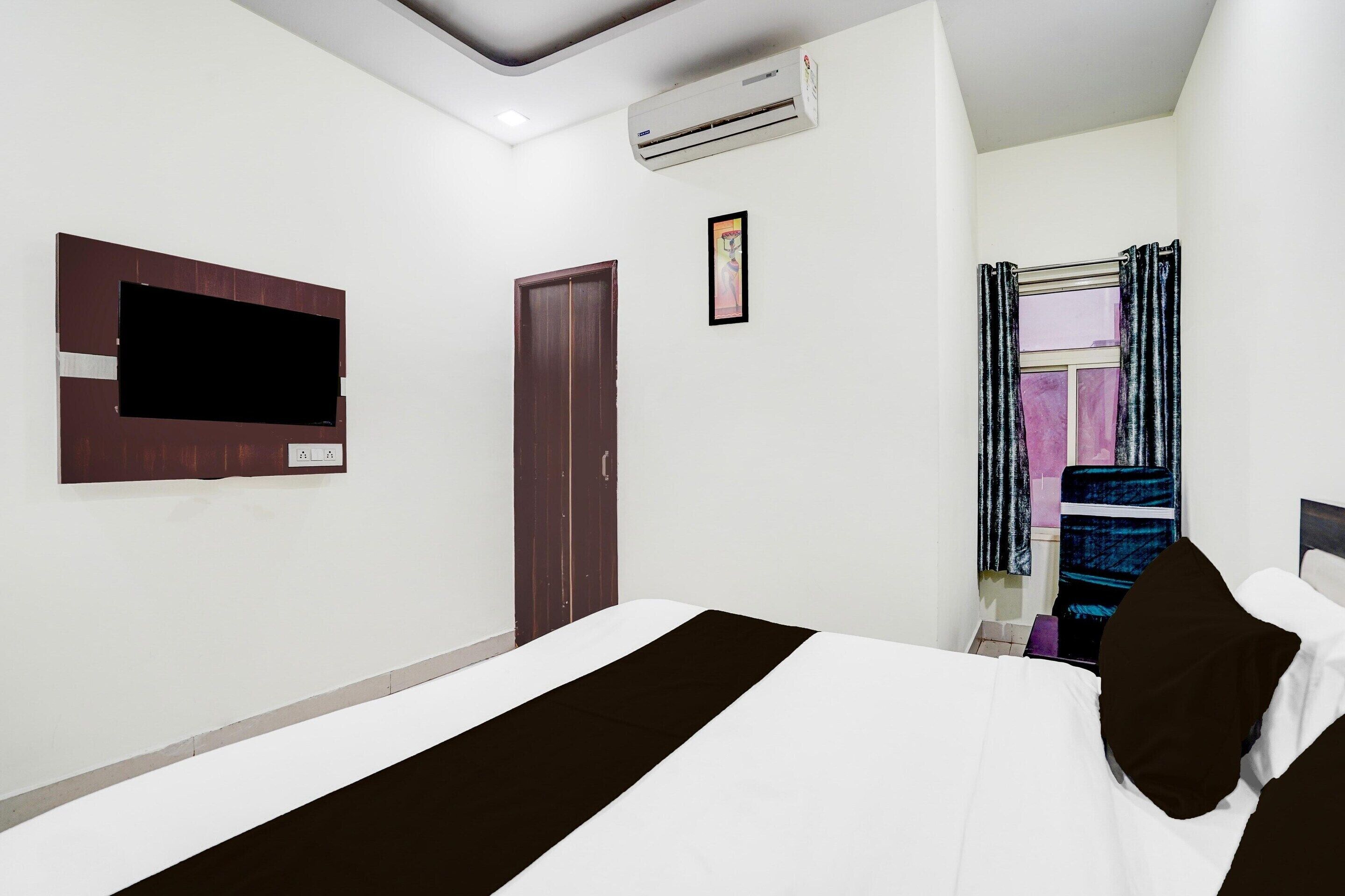 Een hotelkamer met een bed, een flatscreen-tv aan de muur, een airconditioningapparaat, een deur en een raam met gordijnen.