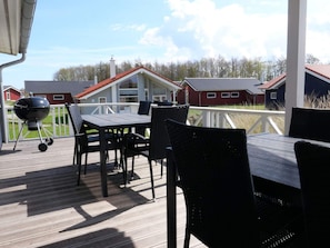 A covered outdoor deck with a table and chairs, a barbecue grill, and a view of red houses in the background. - 4 Star Holiday Home in Grossenbrode (Grossenbrode)