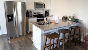 A modern kitchen with a stainless steel refrigerator, microwave, and stove, a granite countertop island with bar stools, and white cabinetry.